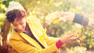 A person wearing a bright yellow coat and a knit hat playfully reaches for autumn leaves being offered by another hand.