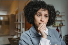 A person with curly hair stands thoughtfully, wearing a grey jacket in a cozy, softly lit indoor setting.