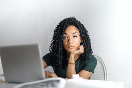 A person with curly hair rests their chin on their hand while looking thoughtfully at a laptop on a white desk.