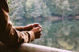 A person's hands clasped on a wooden railing, gazing out at a serene lake surrounded by lush greenery.