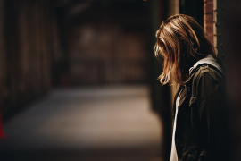 A young woman with shoulder-length hair stands alone, gazing downwards in a dimly lit corridor, creating a mood of contemplation.