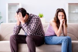 A distressed couple sits apart on a couch, holding their heads in frustration, reflecting a moment of conflict or emotional turmoil.