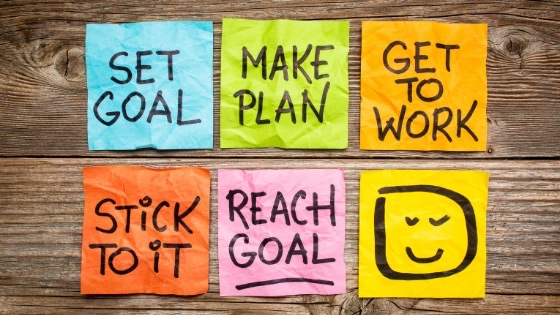 Colorful sticky notes on a wooden surface display motivational phrases: "Set Goal," "Make Plan," "Get to Work," "Stick to It," "Reach Goal," and a smiley face.