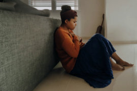 A person sits against a wall, wearing a cozy orange sweater and a long blue skirt, in a serene indoor environment.