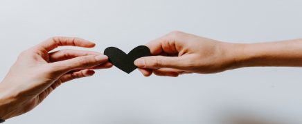 Two hands reaching out to each other, holding a small black paper heart between them against a light background.