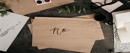 Artistic workspace with brown paper, watercolor paints, green leaves, glasses, and a prominently displayed handwritten "no."