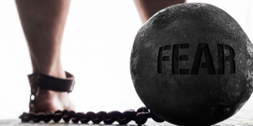 A heavy ball inscribed with "FEAR" is chained to a person's ankle, symbolizing the burden of fear holding them down.