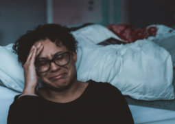 A person sits on a bed, resting their head on their hand, surrounded by crumpled blankets in a dimly lit room.