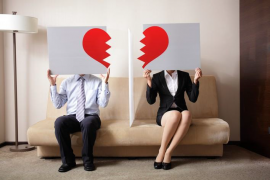 A man and woman sit on a couch holding signs with broken heart graphics, symbolizing a strained or ending relationship.