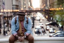 A person in a denim jacket and hat sits on a ledge, gazing over a bustling city street at sunset.