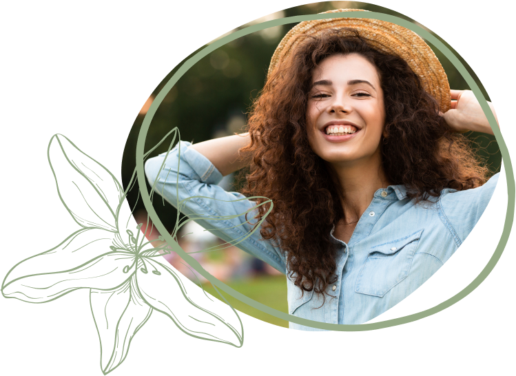 A woman with curly hair in a straw hat poses playfully outdoors, wearing a light blue shirt against a blurred park background.